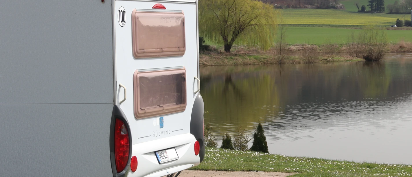 Ein Wohnwagen an einem See, im Hintergrund sieht man Wald auf einem Berg.