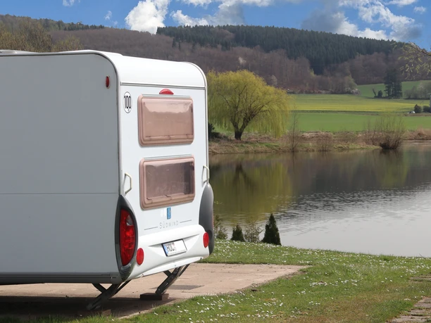 Camp Feuerland Ein Wohnwagen an einem See, im Hintergrund sieht man Wald auf einem Berg.
