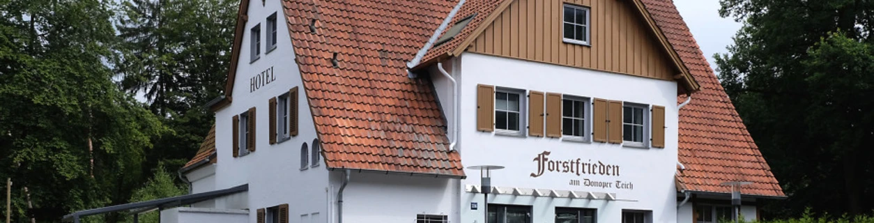 Hotel Forstfrieden, zweistöckiges Gebäude mit roten Dachziegeln, liegt in grüner, bewaldeter Umgebung.