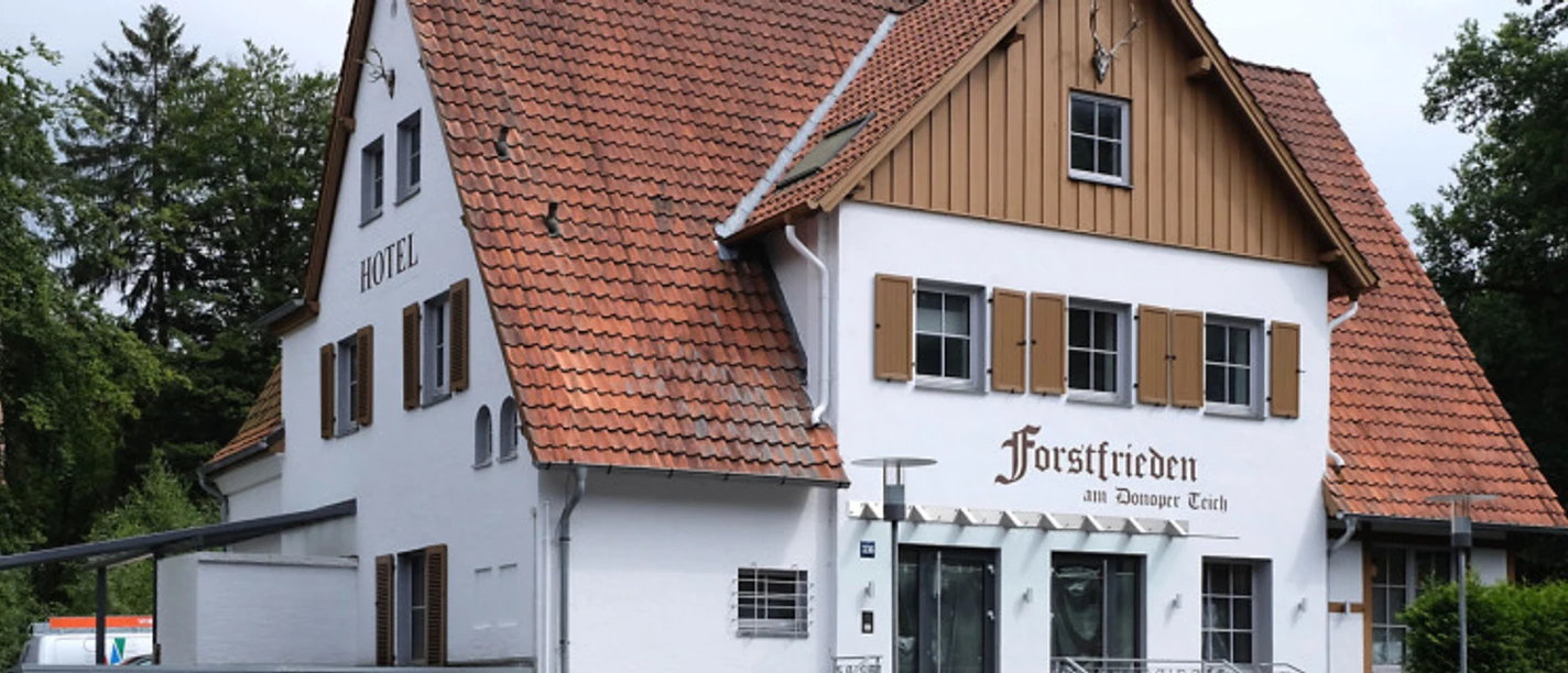 Hotel Forstfrieden, zweistöckiges Gebäude mit roten Dachziegeln, liegt in grüner, bewaldeter Umgebung.