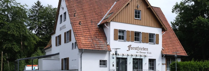 Forstfrieden.jpg Hotel Forstfrieden, zweistöckiges Gebäude mit roten Dachziegeln, liegt in grüner, bewaldeter Umgebung.