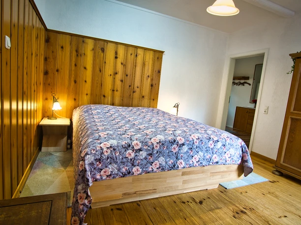 Schlafzimmer mit Doppelbett, floraler Bettdecke, Holzwand und Holzboden.