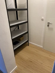 FeWo_Hille-Oberluebbe Schlichter Hausflur mit hellem Laminatboden, leerem Metallregal und weißer Tür in moderner Optik.