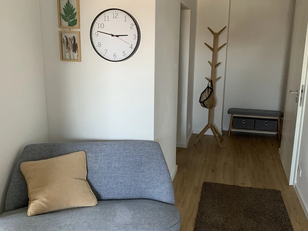 FeWo_Hille-Oberluebbe Flur mit Holzboden, grauem Sofa, Kleiderständer, runder Uhr und Dekobildern an der Wand.