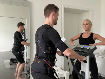 EMS Maximale Erfolge in nur 20 Minuten pro Woche Ein Mann im EMS-Anzug trainiert unter Aufsicht einer Trainerin an einem Fitnessgerät in einem Studio.