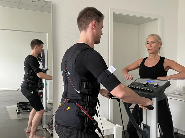 EMS Maximale Erfolge in nur 20 Minuten pro Woche Ein Mann im EMS-Anzug trainiert unter Aufsicht einer Trainerin an einem Fitnessgerät in einem Studio.