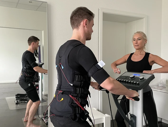 EMS Maximale Erfolge in nur 20 Minuten pro Woche Ein Mann im EMS-Anzug trainiert unter Aufsicht einer Trainerin an einem Fitnessgerät in einem Studio.