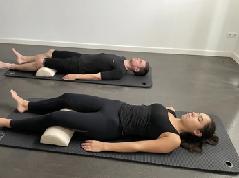 Ganzheitliche Physiotherapie Zwei Personen liegen entspannt auf Matten, vertieft in Entspannung mit Unterstützung von Kissen.