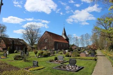 St. Jobst Kirche Odisheim Prächtige St. Jobst Backsteinkirche mit spitzem Turm und grünem Friedhof unter blauem Himmel.