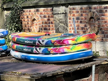 Standup paddling Drei bunte Stand-up-Paddle-Boards liegen gestapelt auf einem Holzdeck vor einer Ziegelmauer.