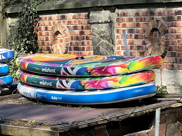 Standup paddling Drei bunte Stand-up-Paddle-Boards liegen gestapelt auf einem Holzdeck vor einer Ziegelmauer.