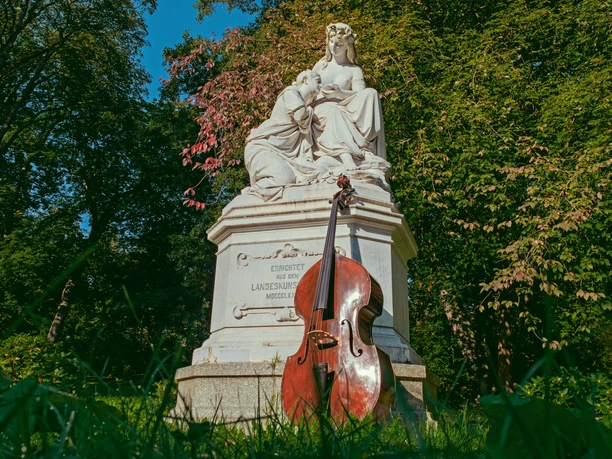 ChursächsischerSommer-Motiv©JanBräuer.jpg Steinskulptur mit Geige im Park, umgeben von üppigem Grün und blauem Himmel.