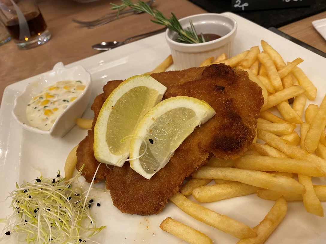 Zwei Zitronenscheiben auf knusprigem Schnitzel, serviert mit Pommes und Kräuterdip, dekoriert mit Sprossen.