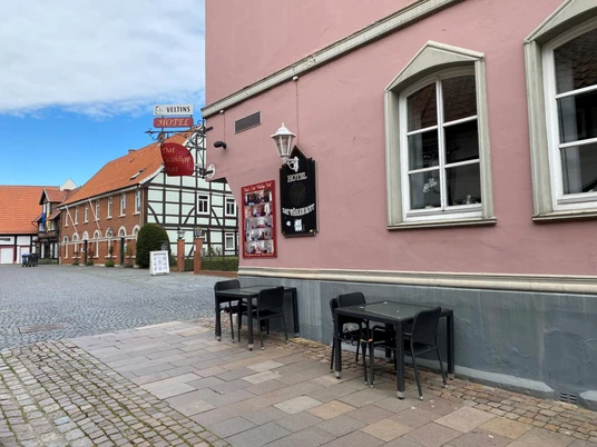 Pup Dat Wählige Rott Ein ruhiger, gepflasterter Platz mit Tischen und Stühlen vor einem rosafarbenen Gebäude im Fachwerkstil.