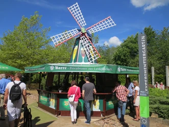 Bier Boulevard Menschen genießen Getränke an einem Bierstand mit Windmühle, umgeben von grünen Bäumen.
