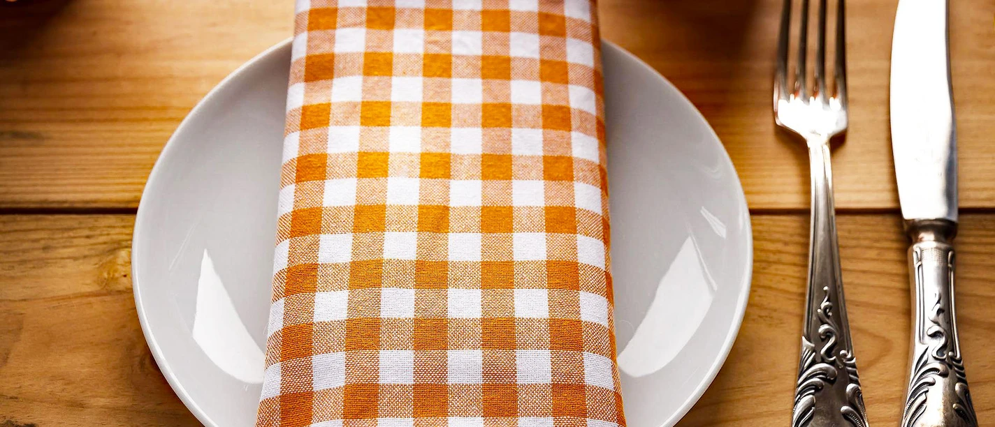 Gastronomie Teller mit karierter Serviette und Besteck auf Holztisch, dazu ein Glas im Hintergrund.