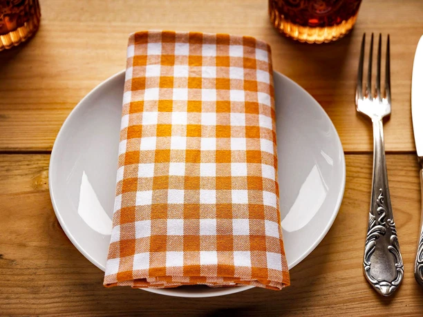 Gastronomie Teller mit karierter Serviette und Besteck auf Holztisch, dazu ein Glas im Hintergrund.