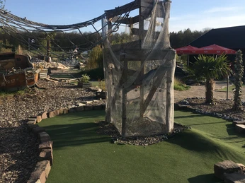 Der Turm im Vordergrund eines Minigolfplatzes ist von Netzen umgeben, im Hintergrund sind Pflanzen.