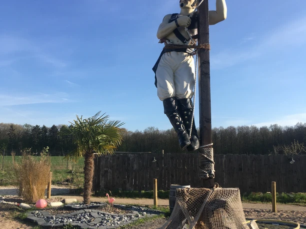 Piratenskulptur im Activity Park Hagenburg, an einem Pfahl angebunden, vor blauem Himmel und Wald.
