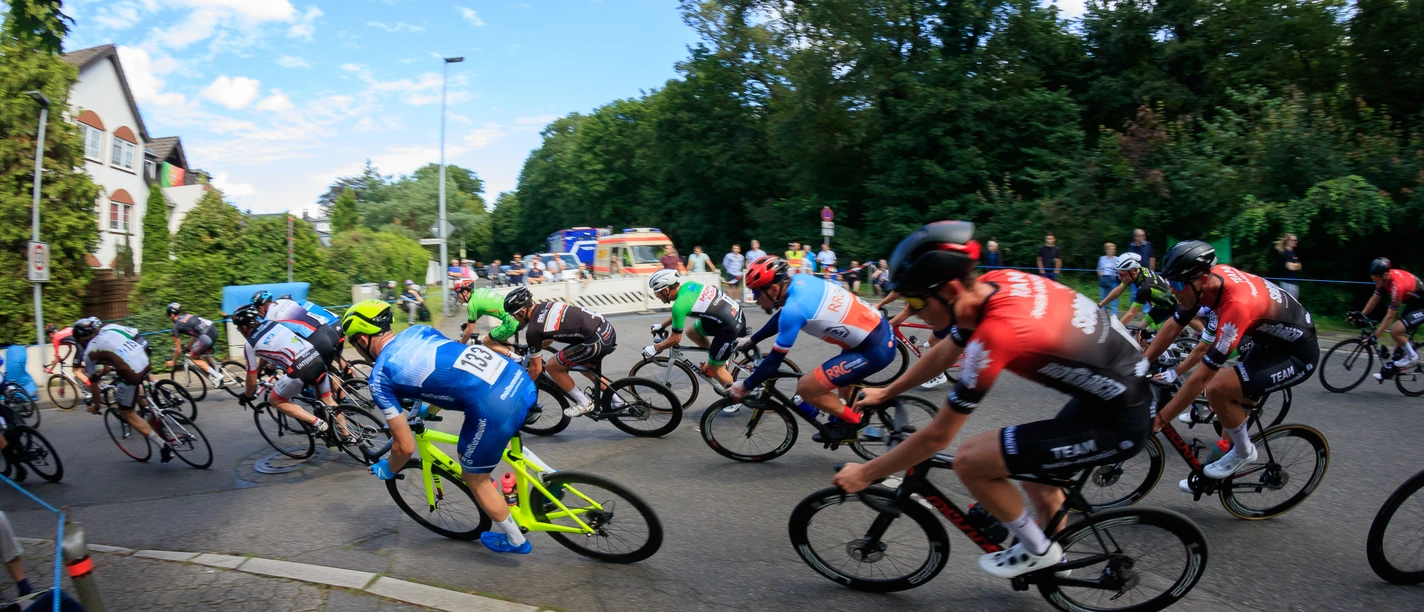 Rennen "Rund in Refrath" Rennradfahrer in einem Wettbewerb rasen eine kurvige Straße entlang, umgeben von Bäumen.