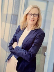 Dr. Dr. med. Susanne Hinze Dr. Susanne Hinze, eine Frau mit blonden Haaren und Brille, steht lächelnd in einem dunkelblauen Blazer.