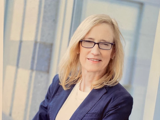 Dr. Dr. med. Susanne Hinze Dr. Susanne Hinze, eine Frau mit blonden Haaren und Brille, steht lächelnd in einem dunkelblauen Blazer.