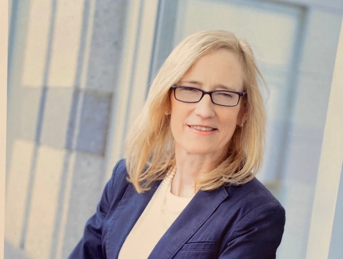 Dr. Dr. med. Susanne Hinze Dr. Susanne Hinze, eine Frau mit blonden Haaren und Brille, steht lächelnd in einem dunkelblauen Blazer.