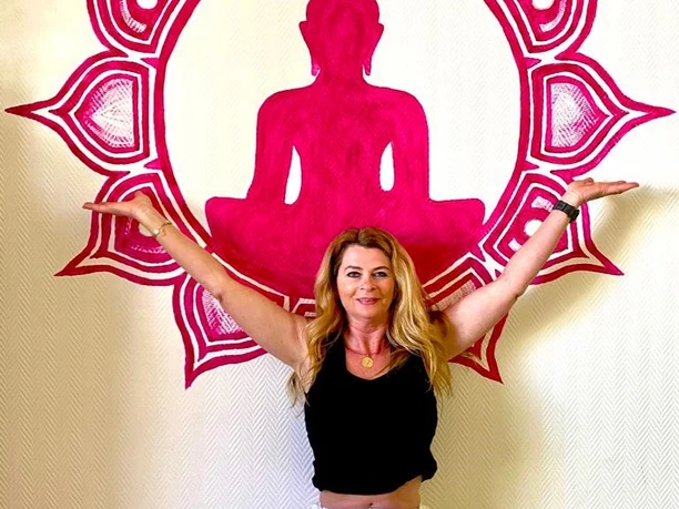 Heidi Lödige Yoga Eine Frau in weißer Hose und schwarzem Shirt meditiert vor einer pinken Buddha-Silhouette an der Wand.