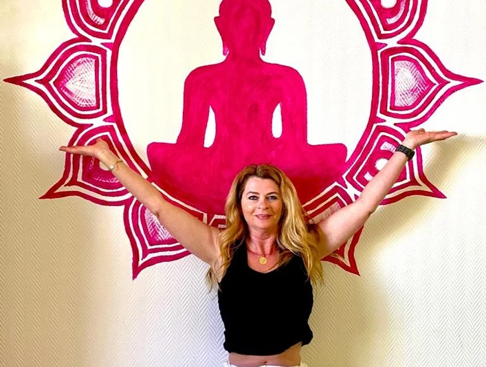 Heidi Lödige Yoga Eine Frau in weißer Hose und schwarzem Shirt meditiert vor einer pinken Buddha-Silhouette an der Wand.