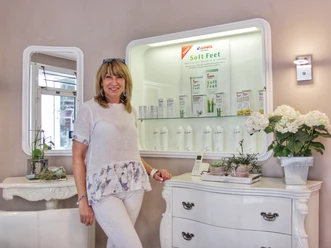 Elena Lang Eine Frau in weißer Kleidung steht lächelnd vor einer Vitrine mit Fußpflegeprodukten in einem Salon.