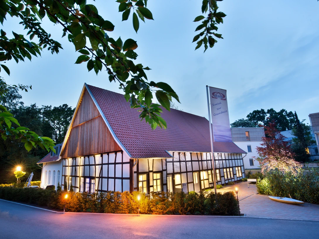 Nachtaufnahme mit Baum Fachwerkhaus bei Dämmerung, beleuchtet mit Laternen, umgeben von Bäumen und einem klaren Himmel.