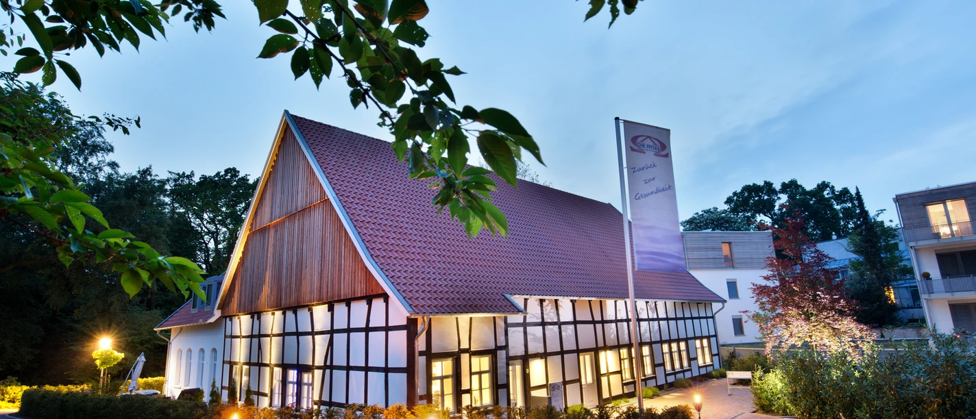 Nachtaufnahme mit Baum Fachwerkhaus bei Dämmerung, beleuchtet mit Laternen, umgeben von Bäumen und einem klaren Himmel.