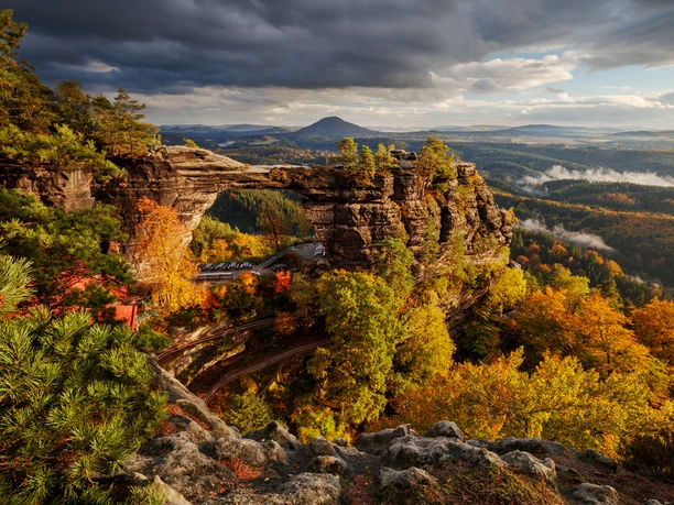 Prebischtor Felsformation mit natürlichem Bogen in herbstlicher Landschaft, umgeben von buntem Laub und dunklen Wolken am Himmel, im Hintergrund Hügel.