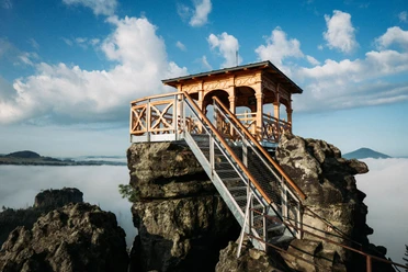 Dittersbacher Felsen Holzpavillon auf einem Felsen mit Treppe, umgeben von Nebel und blauem Himmel mit Wolken; ruhige, malerische Atmosphäre.