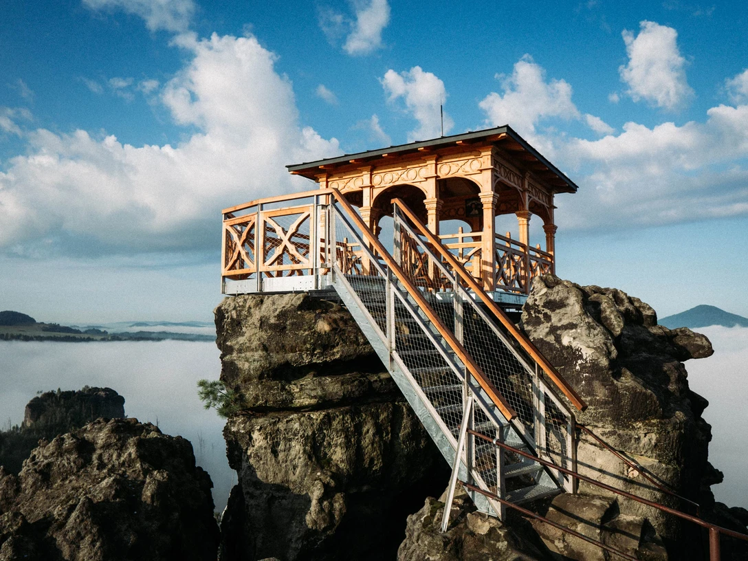 Dittersbacher Felsen Holzpavillon auf einem Felsen mit Treppe, umgeben von Nebel und blauem Himmel mit Wolken; ruhige, malerische Atmosphäre.