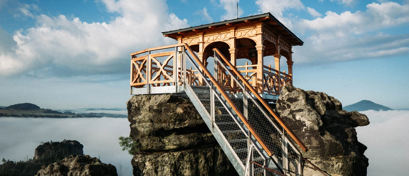 Dittersbacher Felsen Holzpavillon auf einem Felsen mit Treppe, umgeben von Nebel und blauem Himmel mit Wolken; ruhige, malerische Atmosphäre.