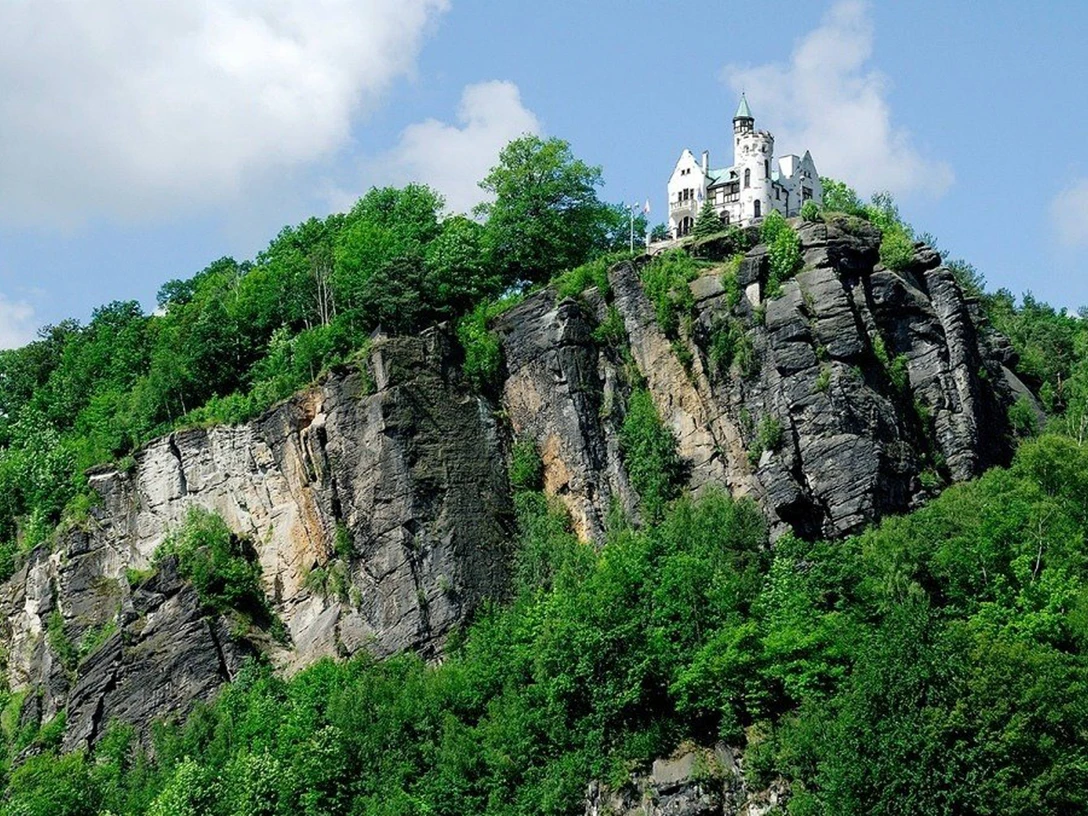 Klettersteig Děčín Ein weißes Schloss thront auf einem bewaldeten Felsen unter blauem Himmel mit Wolken, umgeben von üppigem Grün.A white castle sits enthroned on a wooded rock under a blue sky with clouds, surrounded by lush greenery.Bílý hrad trůní na zalesněné skále pod modrou oblohou s mraky, obklopen bujnou zelení.Biały zamek stoi na zalesionej skale pod błękitnym niebem z chmurami, otoczony bujną zielenią.Een wit kasteel troont op een beboste rots onder een blauwe hemel met wolken, omringd door weelderig groen.Un castello bianco troneggia su una roccia boscosa sotto un cielo blu con nuvole, circondato da una vegetazione lussureggiante.