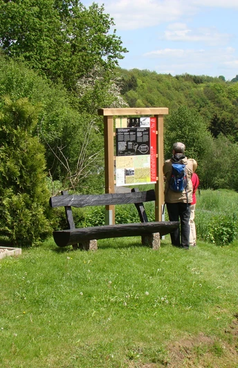 Infotafel am Böllweg Zwei Personen lesen eine Infotafel in einer grünen, hügeligen Landschaft mit Bäumen und Bänken.