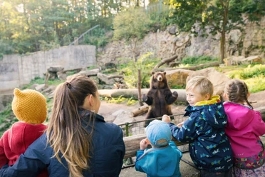 Zoo Děčín Eine Gruppe von Kindern und ein Erwachsener beobachten einen stehenden Bären in einem Zoo-Gehege, umgeben von Bäumen und Felsen.