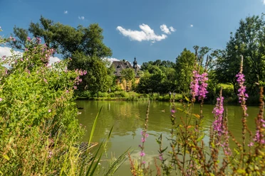 Blick auf Burg Zweiffel in Herrenstrunden Historisches Schloss mit Turm umgeben von grünen Bäumen und einem Teich im Vordergrund.