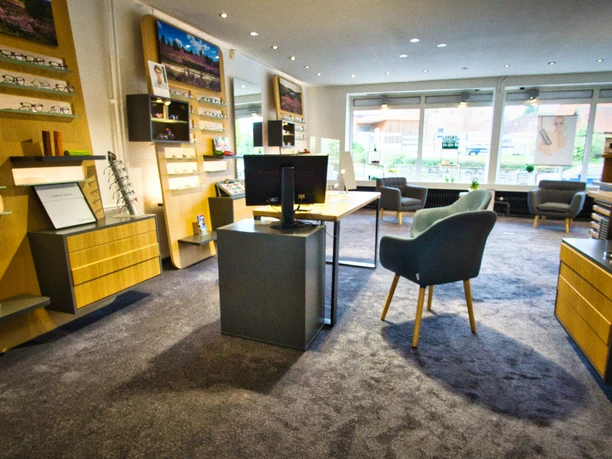 Schröder Optik & Kurzhals GmbH Helles Optikergeschäft mit Brillenregalen aus Holz, grauen Sesseln und großen Schaufenstern.