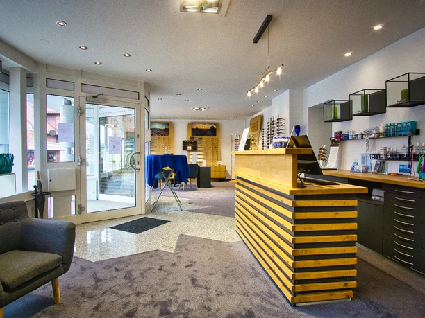 Innenansicht-Verkaufsbereich Heller Optikerladen mit großer Holztheke, grauem Sessel und Brillenregalen an den Wänden.