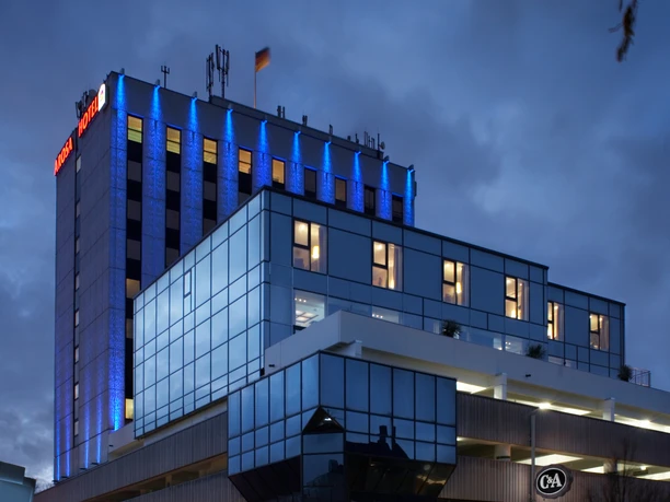 Modernes Hotelgebäude mit blauer Glasfassade und beleuchtetem Dach bei bewölktem Abendhimmel.