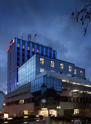 Best Western Plus Arosa Hotel Modernes Hotelgebäude mit blauer Glasfassade und beleuchtetem Dach bei bewölktem Abendhimmel.