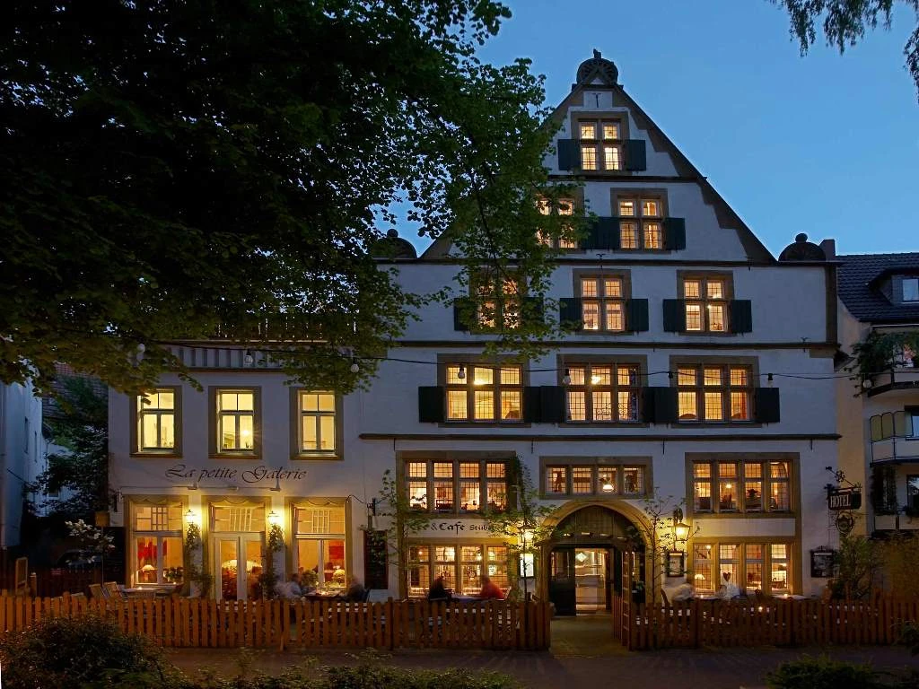 Fachwerkhaus mit leuchtenden Fenstern am Abend, umgeben von Bäumen und Holzzaun, lädt zum Verweilen ein.