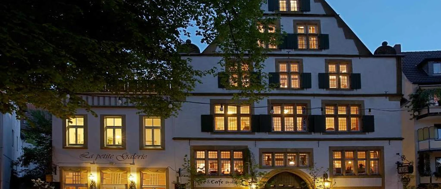 Galerie-Hotel Abdinghof Fachwerkhaus mit leuchtenden Fenstern am Abend, umgeben von Bäumen und Holzzaun, lädt zum Verweilen ein.