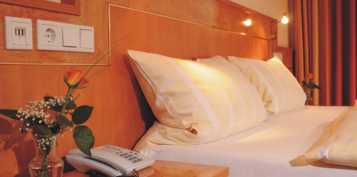 Hotel Heide Residenz Doppelbett mit weißen Kissen, Telefon auf dem Nachttisch, Vase mit Rose im Vordergrund.