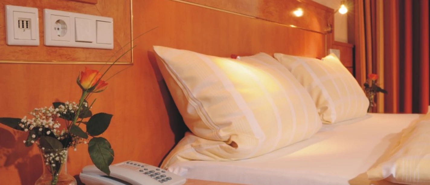 Hotel Heide Residenz Doppelbett mit weißen Kissen, Telefon auf dem Nachttisch, Vase mit Rose im Vordergrund.
