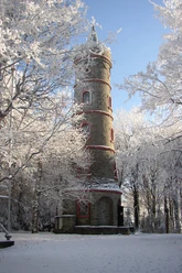 Tannenberg Aussichtsturm Ein runder Steinturm mit roten Akzenten steht in einer verschneiten Winterlandschaft, umgeben von schneebedeckten Bäumen unter klarem, blauem Himmel.