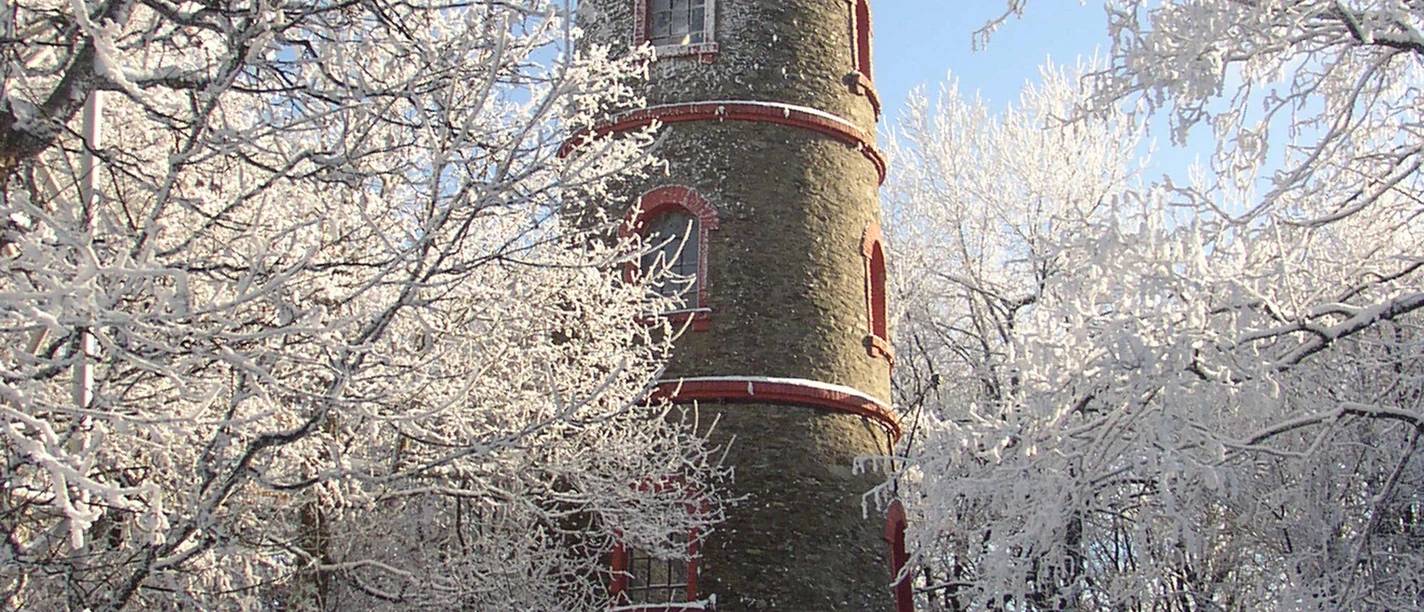 Tannenberg Aussichtsturm Ein runder Steinturm mit roten Akzenten steht in einer verschneiten Winterlandschaft, umgeben von schneebedeckten Bäumen unter klarem, blauem Himmel.